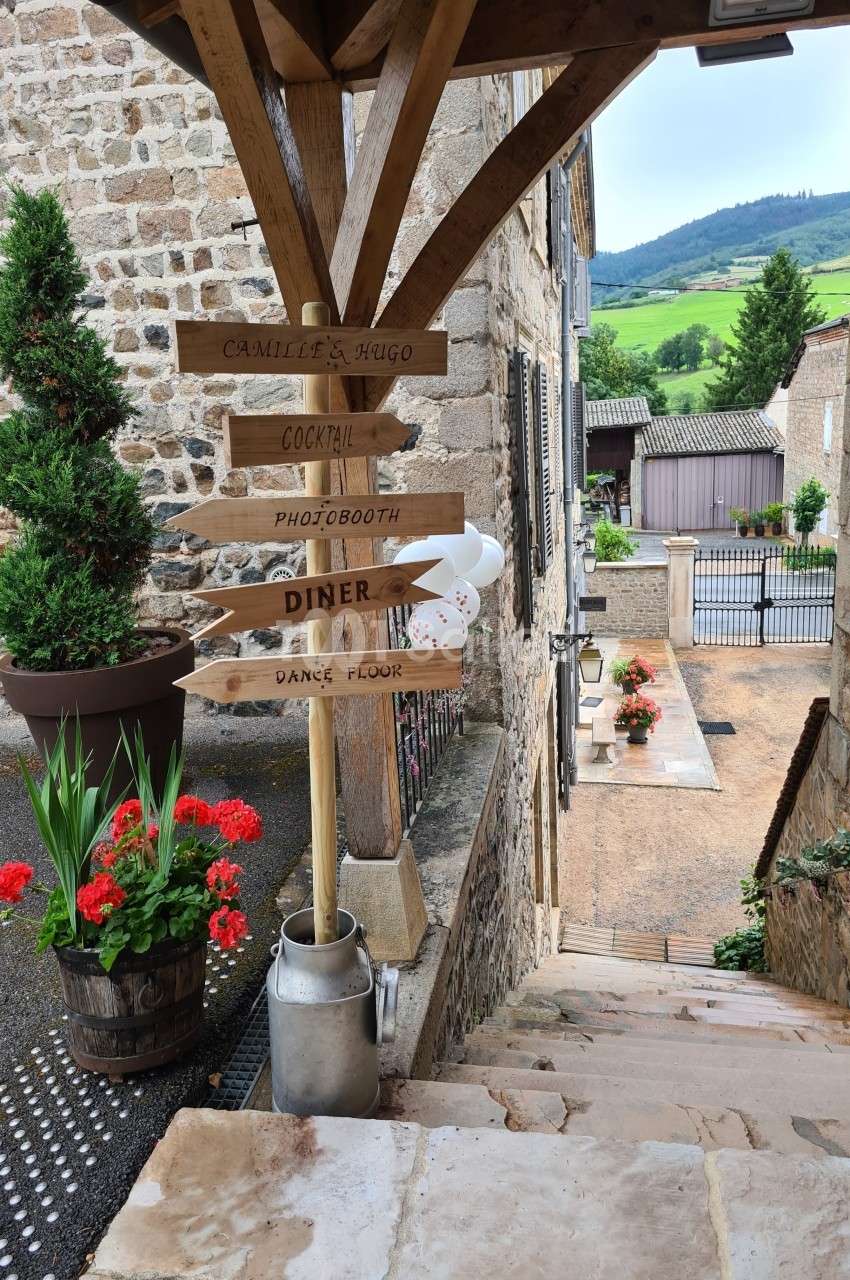Panneau en bois indiquant des directions pour un événement, dans une ruelle pavée bordée de fleurs et de bâtiments en pierre.