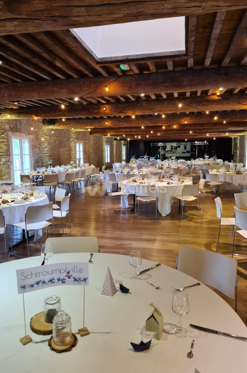 Salle de réception décorée avec des tables rondes dressées, éclairée par des guirlandes lumineuses et des fenêtres en pierre.