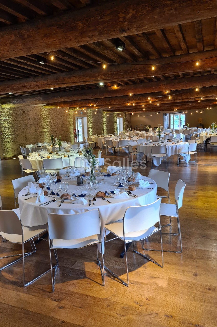 Salle de réception avec tables rondes dressées, nappes blanches et chaises modernes, éclairée par des lumières chaleureuses.