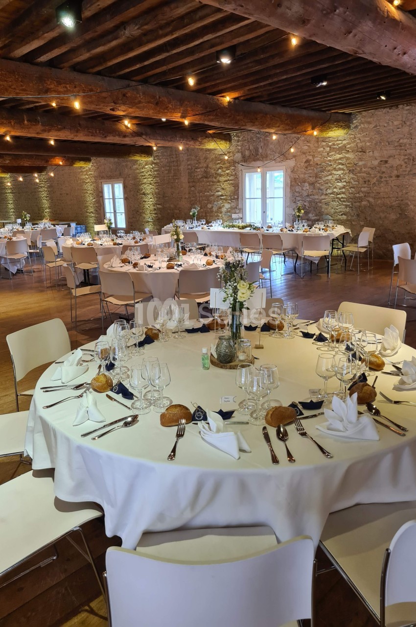 Salle de réception avec tables rondes dressées, nappes blanches, décorations florales et lumière tamisée.