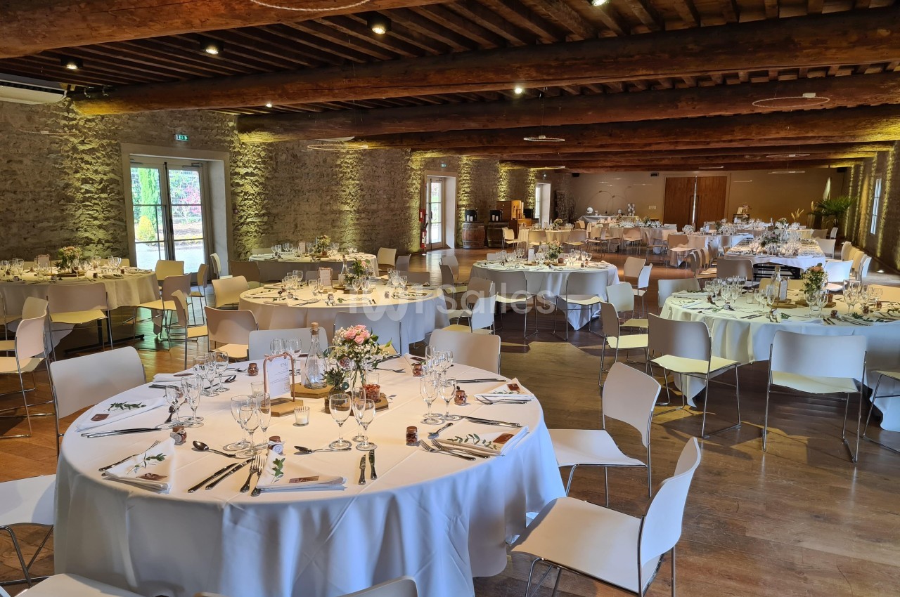 Salle de réception décorée avec des tables rondes dressées pour un événement, dans un cadre en pierre et bois.