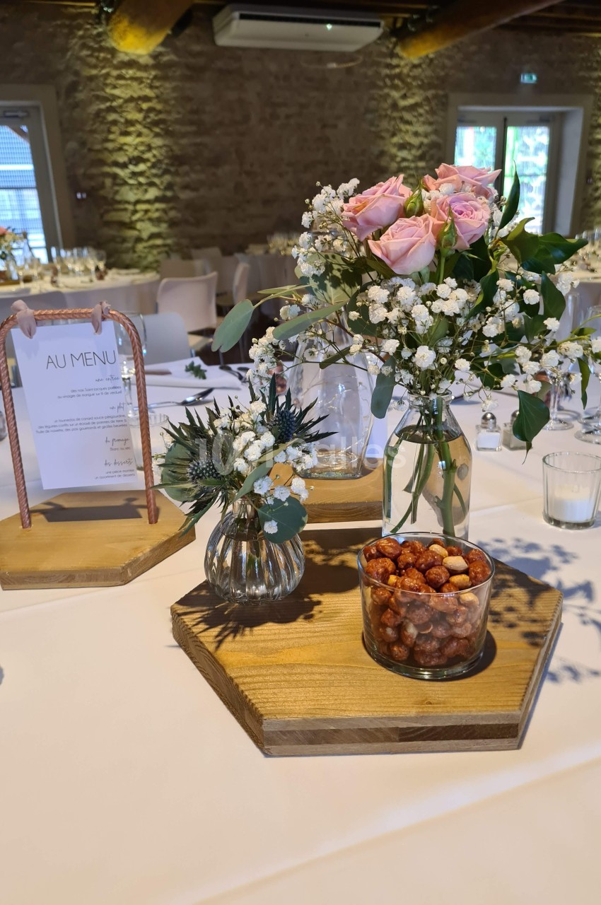 Centre de table avec fleurs roses, gypsophile, pots de fruits secs et menu dans une salle en pierre lumineuse.