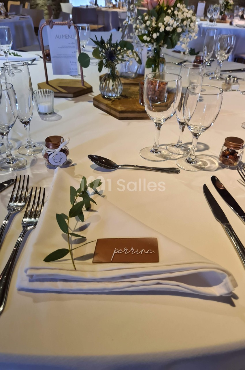 Table dressée pour un repas avec nappe blanche, couverts disposés, marque-place en bois et décoration florale.