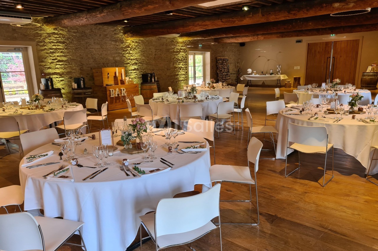 Salle de réception décorée pour un événement, avec tables rondes dressées, chaises blanches et éclairage chaleureux.