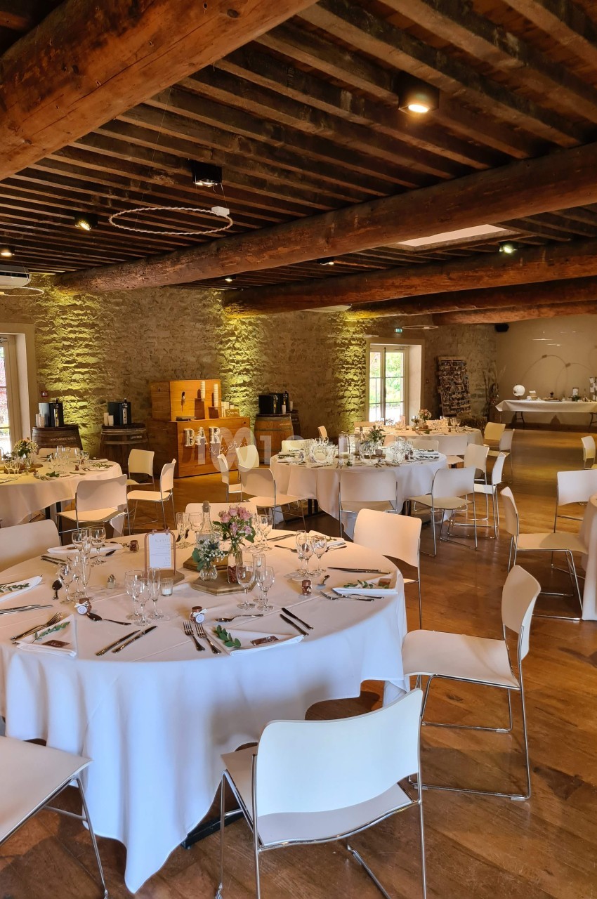 Salle de réception avec tables rondes dressées, chaises blanches et décor rustique en bois et pierre apparente.