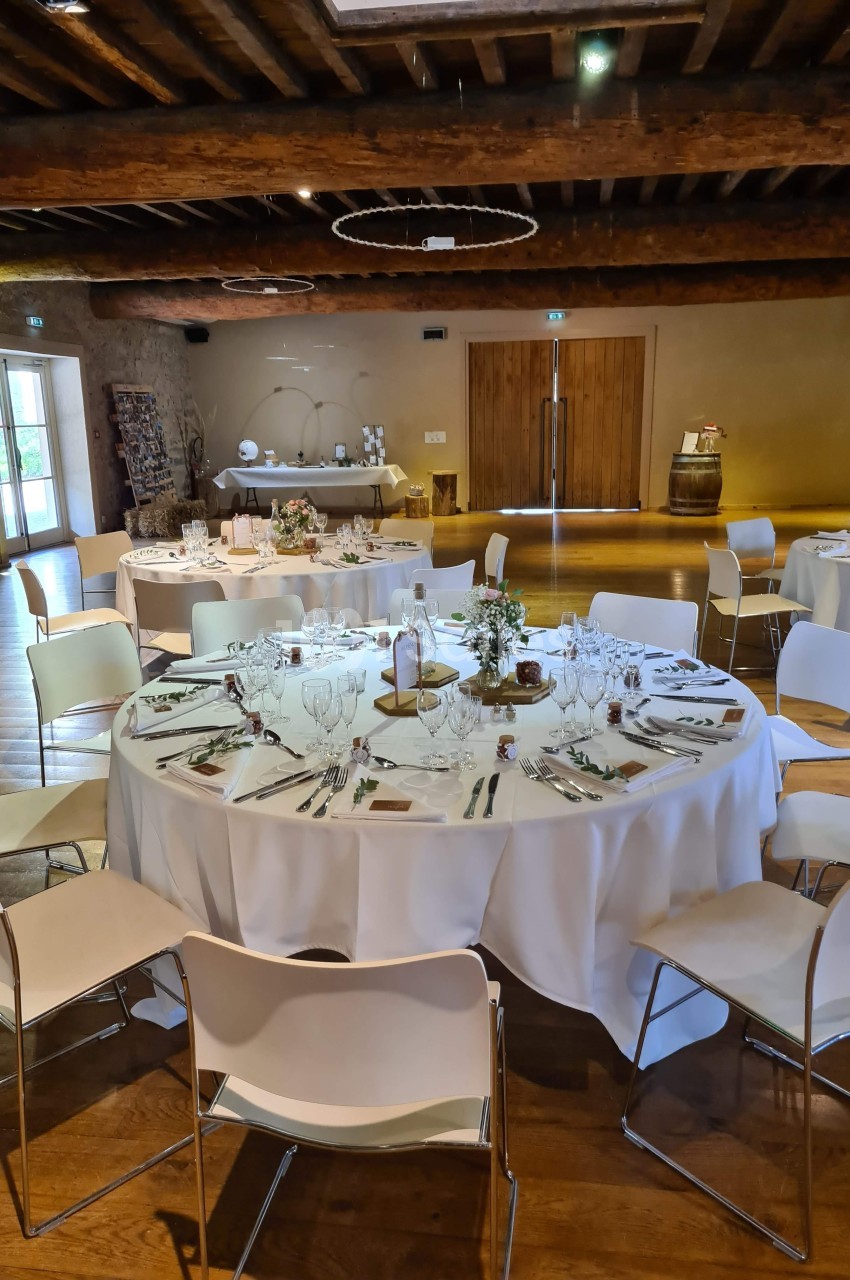 Salle de réception avec tables rondes dressées, nappes blanches, chaises modernes et décoration rustique en bois.