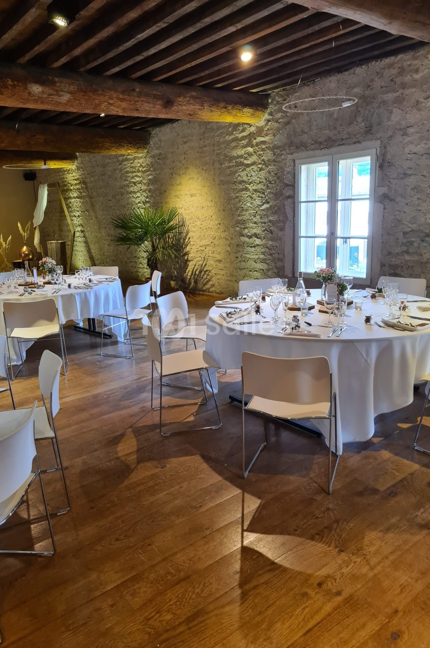 Salle de réception avec murs en pierre, tables rondes dressées et chaises blanches sur un parquet en bois.
