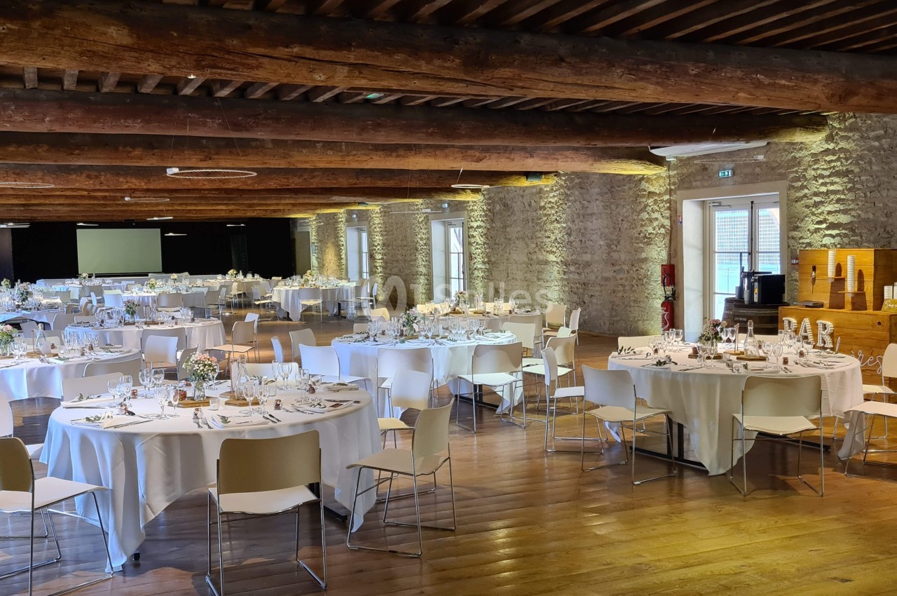 Salle de réception avec tables rondes dressées, chaises blanches, murs en pierre et poutres en bois apparentes.