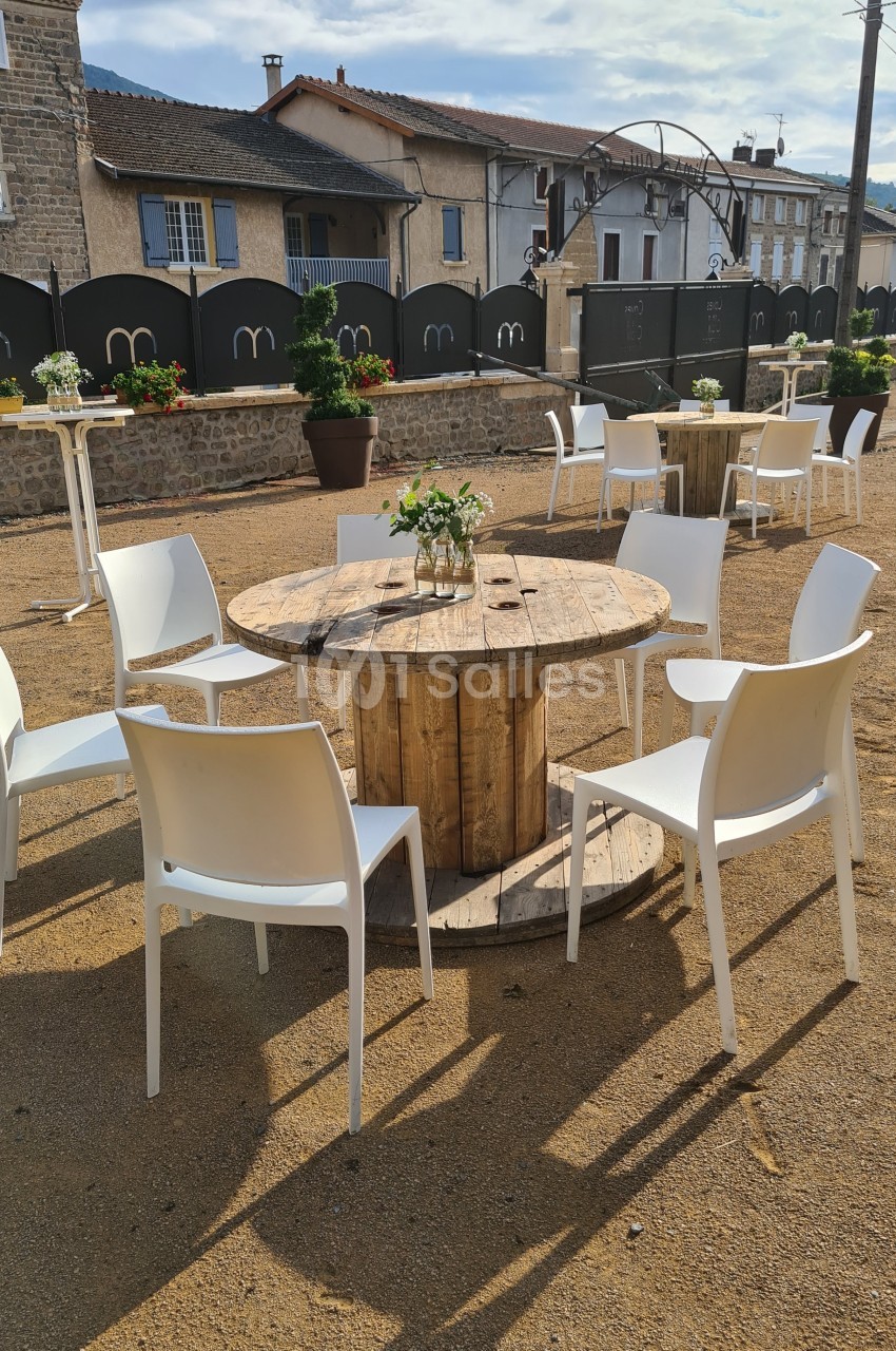 Table ronde en bois de récupération entourée de chaises blanches sur une place extérieure ensoleillée.