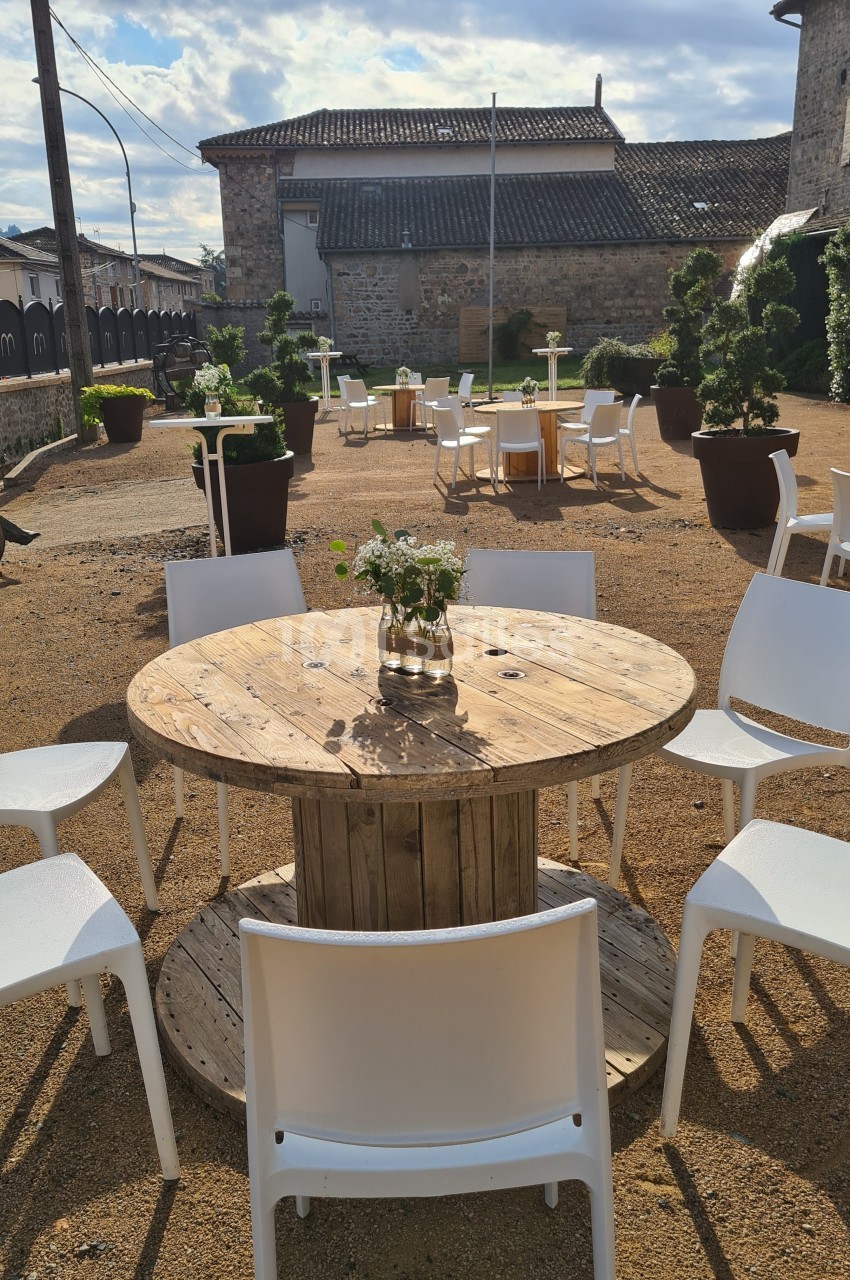 Table ronde en bois entourée de chaises blanches sur une terrasse extérieure en gravier, avec des tables similaires en…