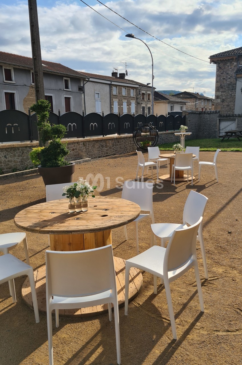 Tables rondes en bois avec chaises blanches disposées en extérieur sur un sol en gravier, entourées de bâtiments.