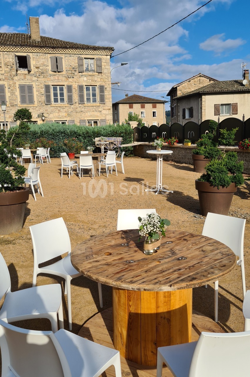 Table ronde en bois entourée de chaises blanches sur une terrasse extérieure avec des bâtiments en pierre en arrière-plan.