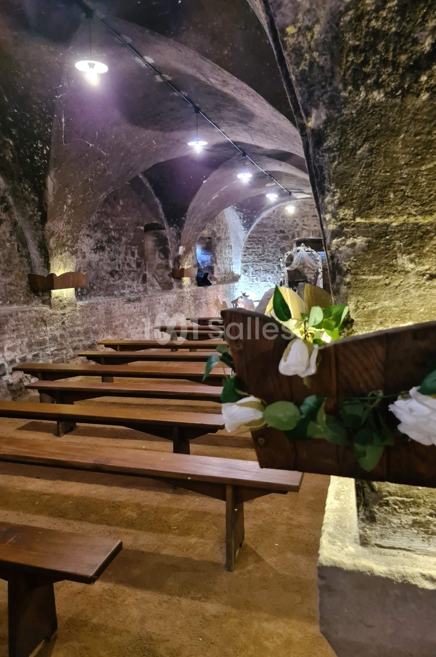 Bancs en bois alignés dans une cave voûtée en pierre, décorée de fleurs blanches et éclairée par des lampes suspendues.