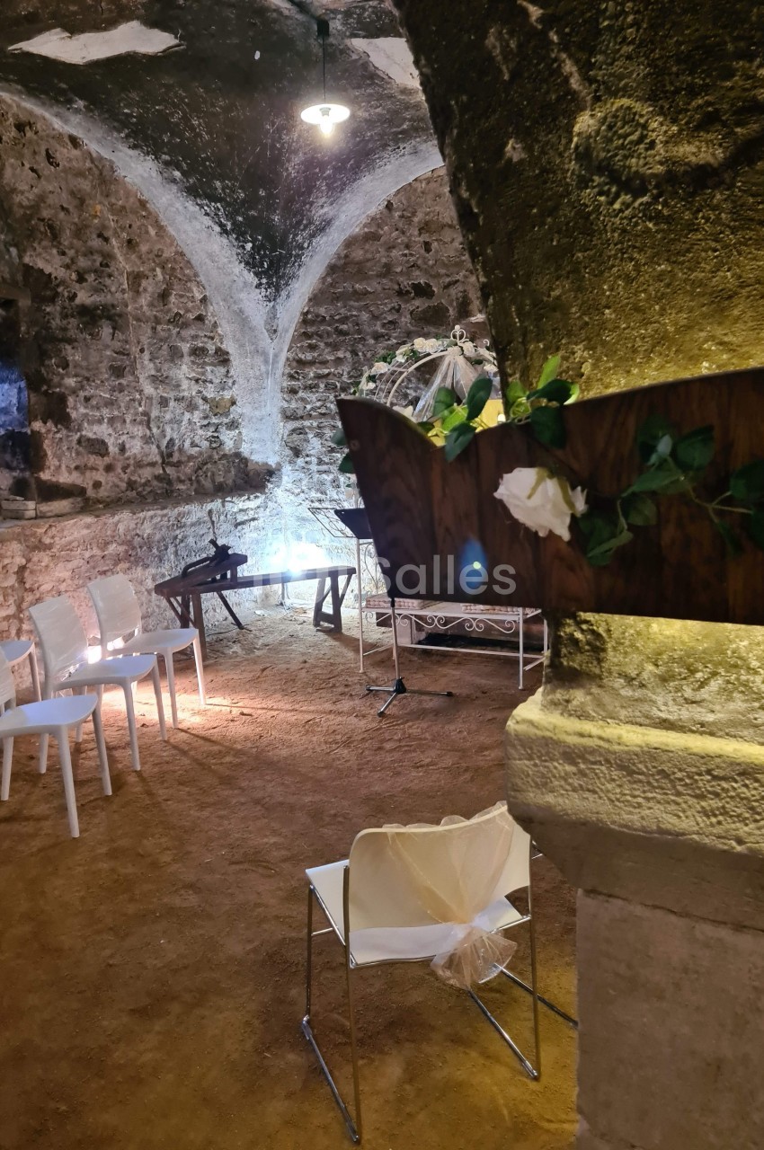 Salle voûtée en pierre avec des chaises blanches disposées pour un événement, décorée de fleurs et éclairée doucement.