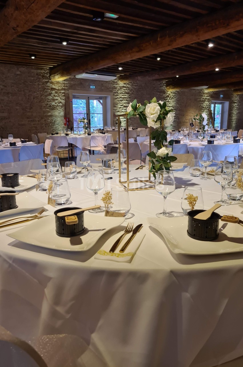 Salle de réception avec tables rondes dressées, nappes blanches, décorations florales et lumière naturelle.