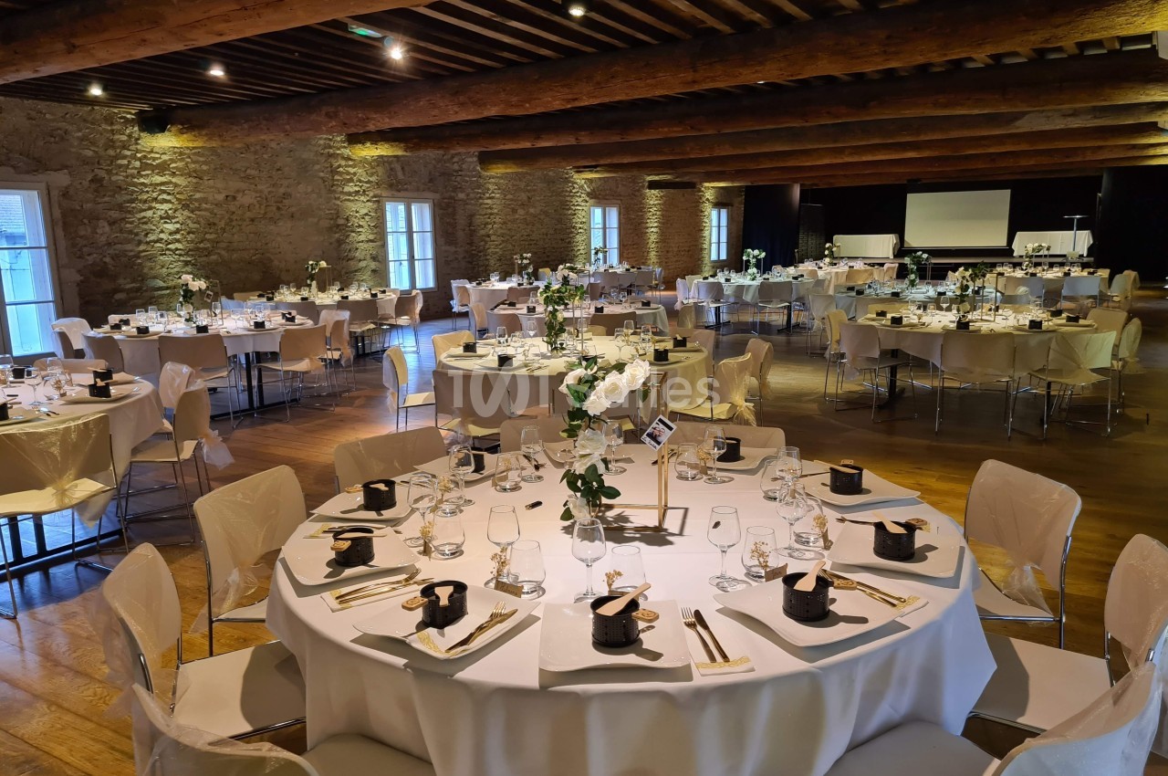 Salle de réception avec tables rondes dressées, nappes blanches, décorations florales et éclairage chaleureux.
