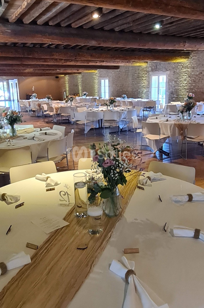 Salle de réception décorée avec des tables rondes, nappes blanches, centres de table fleuris et poutres apparentes au…