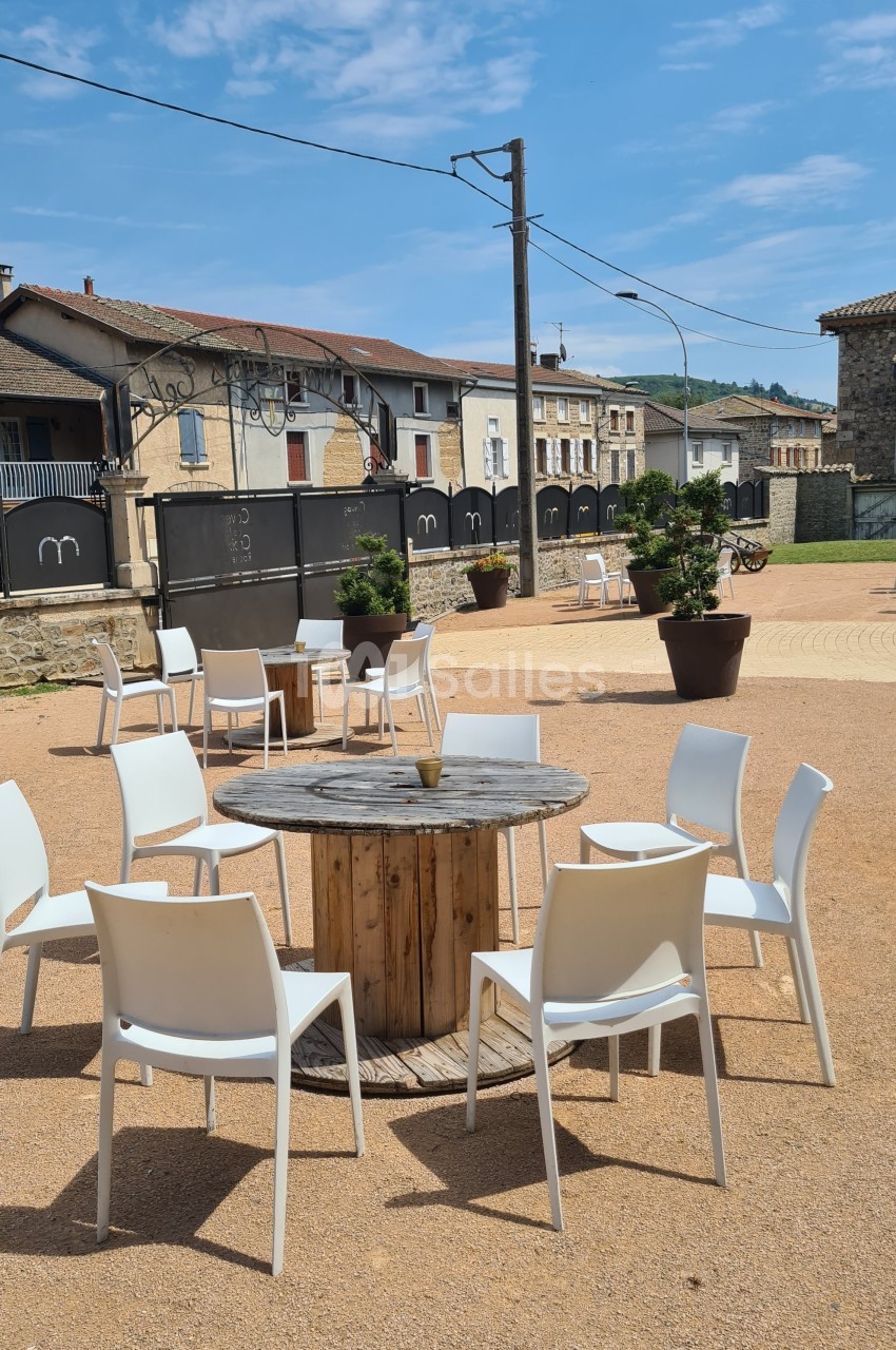 Chaises blanches disposées autour d'une table en bois sur une place ensoleillée avec des bâtiments en arrière-plan.
