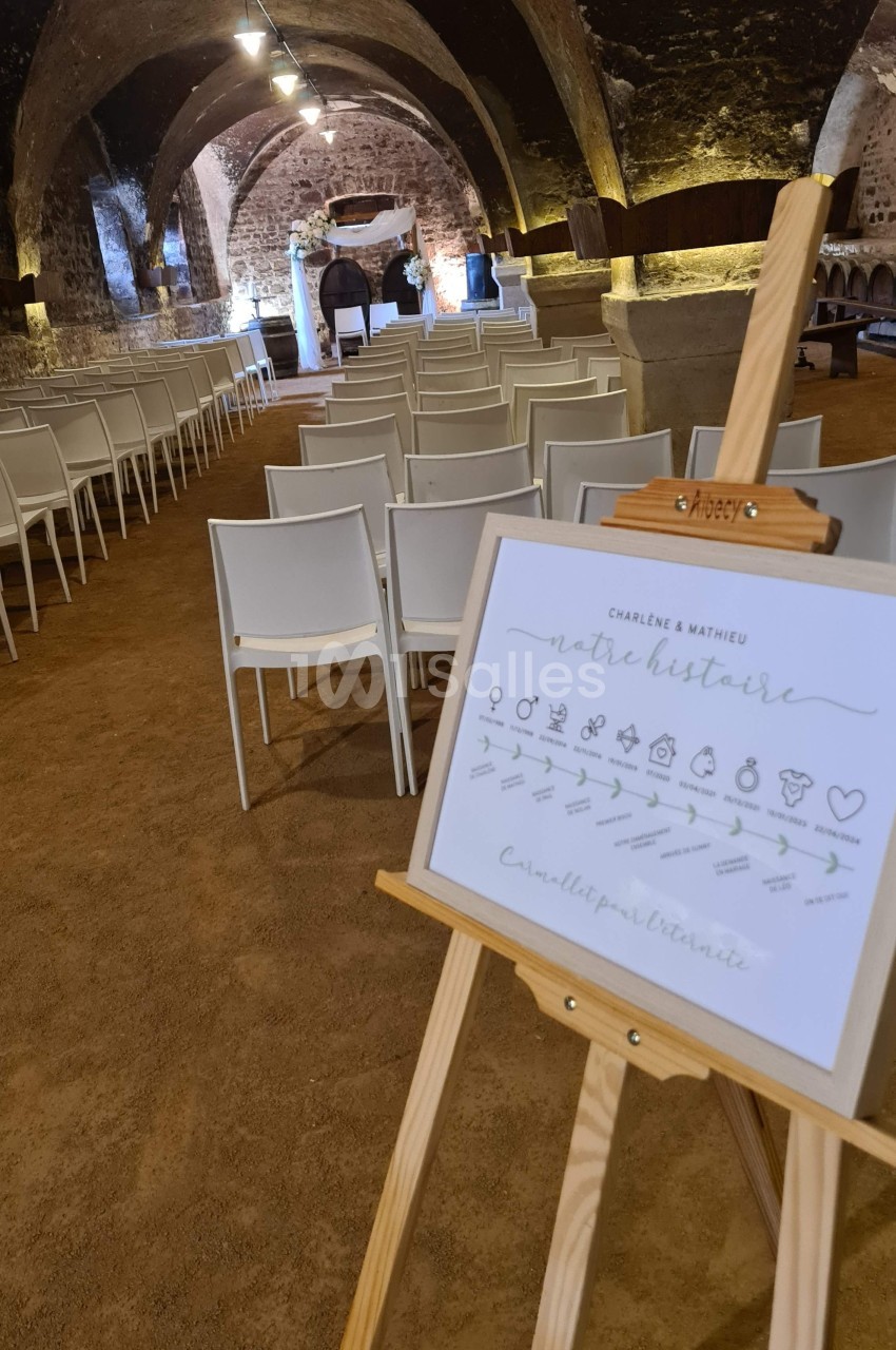 Salle voûtée avec des chaises alignées pour une cérémonie, tableau sur chevalet présentant un programme ou une histoire.