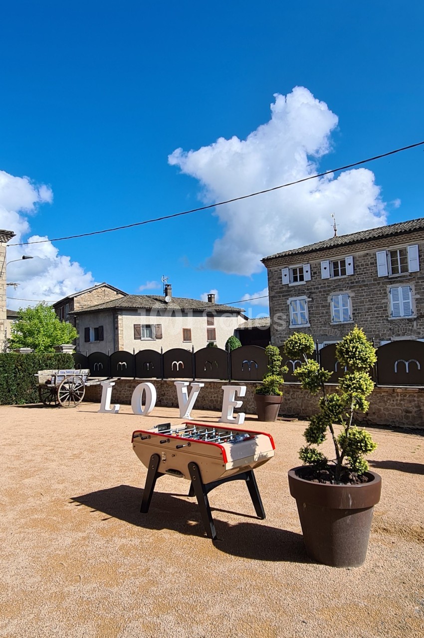 Table de baby-foot sur une place ensoleillée, entourée de pots de plantes, avec des lettres formant ’LOVE’ en arrière-plan.