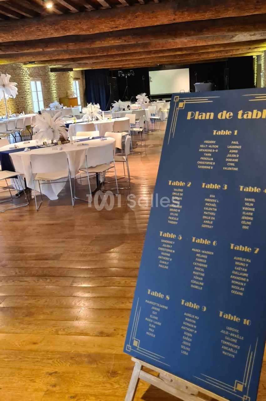 Plan de table sur un chevalet dans une salle de réception avec des tables rondes décorées et un parquet en bois.