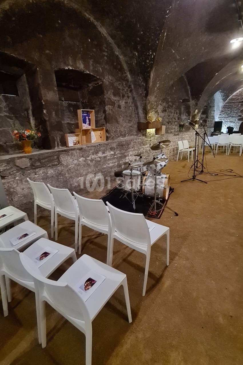 Salle voûtée en pierre avec chaises blanches alignées, une batterie, des micros et des décorations florales.