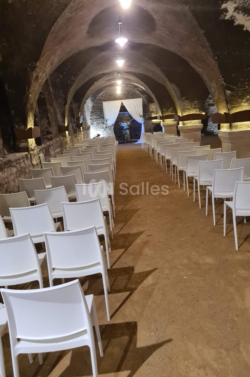 Rangées de chaises blanches alignées dans une salle voûtée en pierre avec un sol en terre battue.