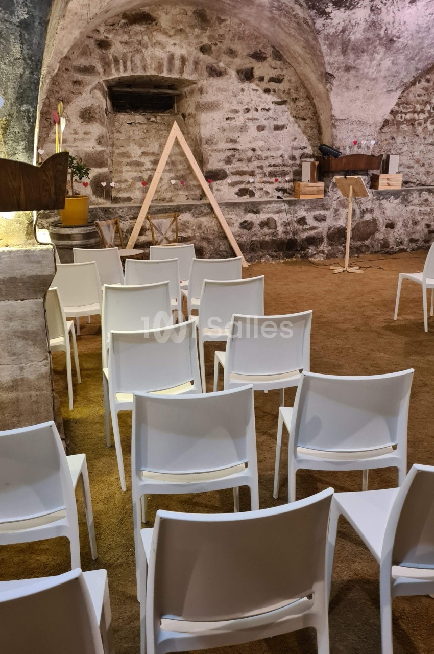 Chaises blanches alignées dans une salle voûtée en pierre avec un présentoir et une structure triangulaire en bois.
