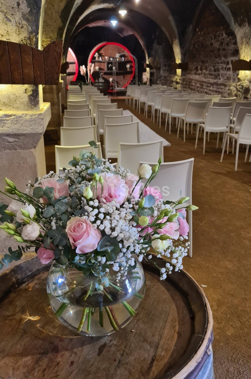 Salle voûtée avec des rangées de chaises blanches, un bouquet de fleurs roses posé sur un tonneau au premier plan.