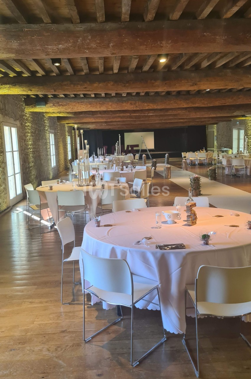 Salle de réception avec tables rondes décorées, chaises blanches et poutres apparentes au plafond.