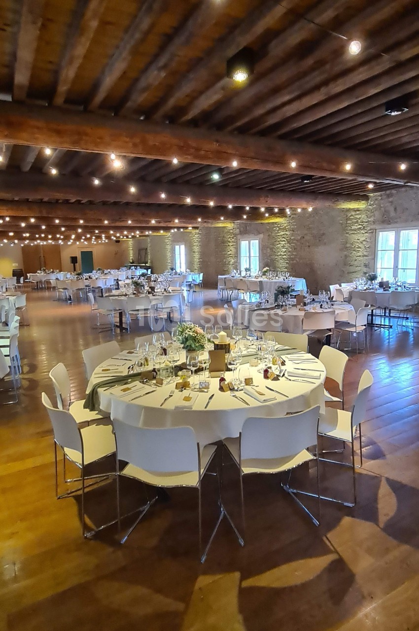 Salle de réception décorée avec des tables rondes dressées, chaises blanches et éclairage chaleureux sous un plafond en bois.