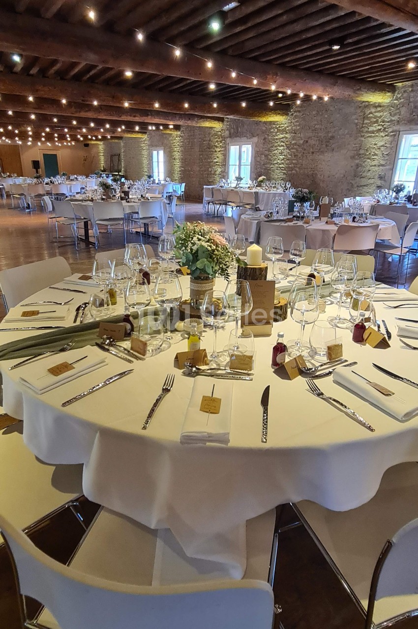 Salle de réception avec tables rondes dressées, nappes blanches, décorations florales et éclairage chaleureux.