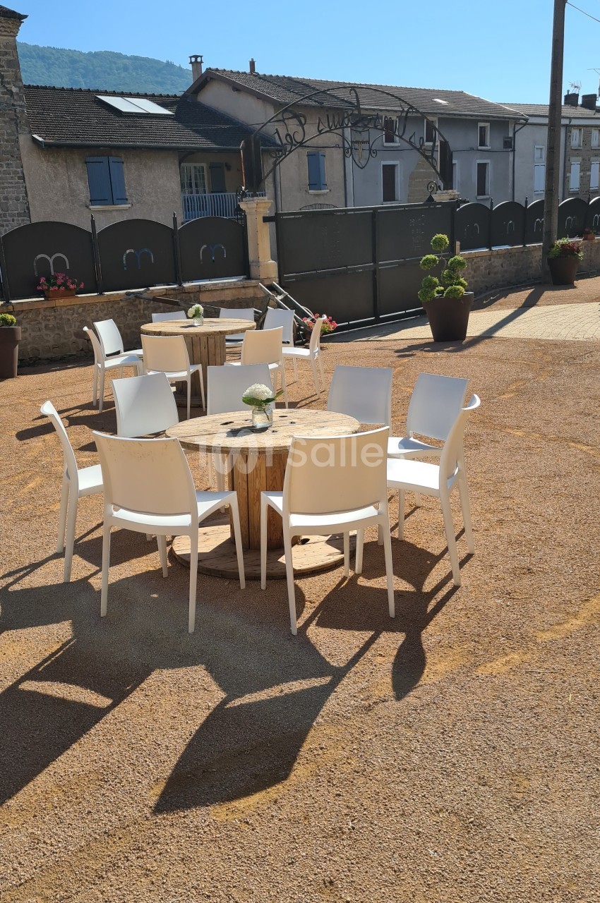 Chaises blanches disposées autour de tables en bois sur une terrasse extérieure en gravier, avec des bâtiments en arrière…