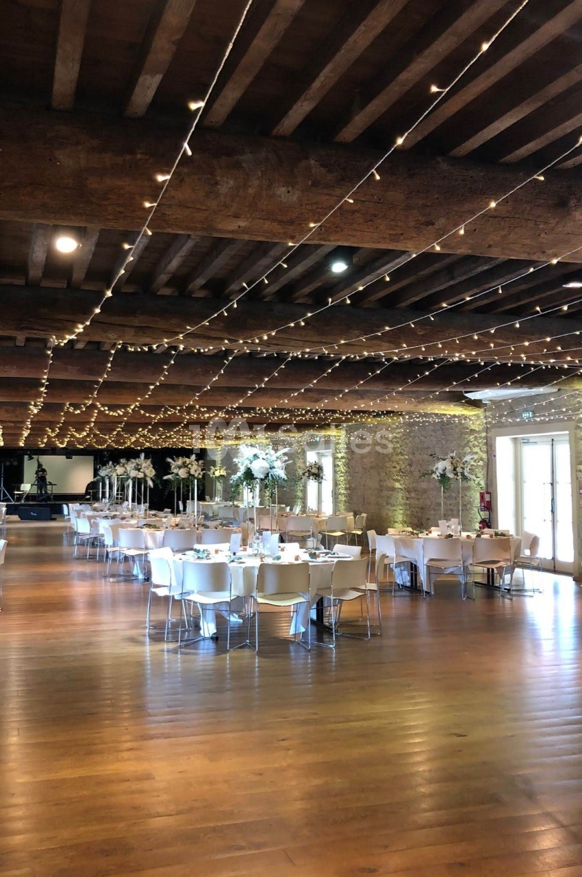 Salle de réception décorée avec des guirlandes lumineuses, tables dressées et poutres apparentes en bois.