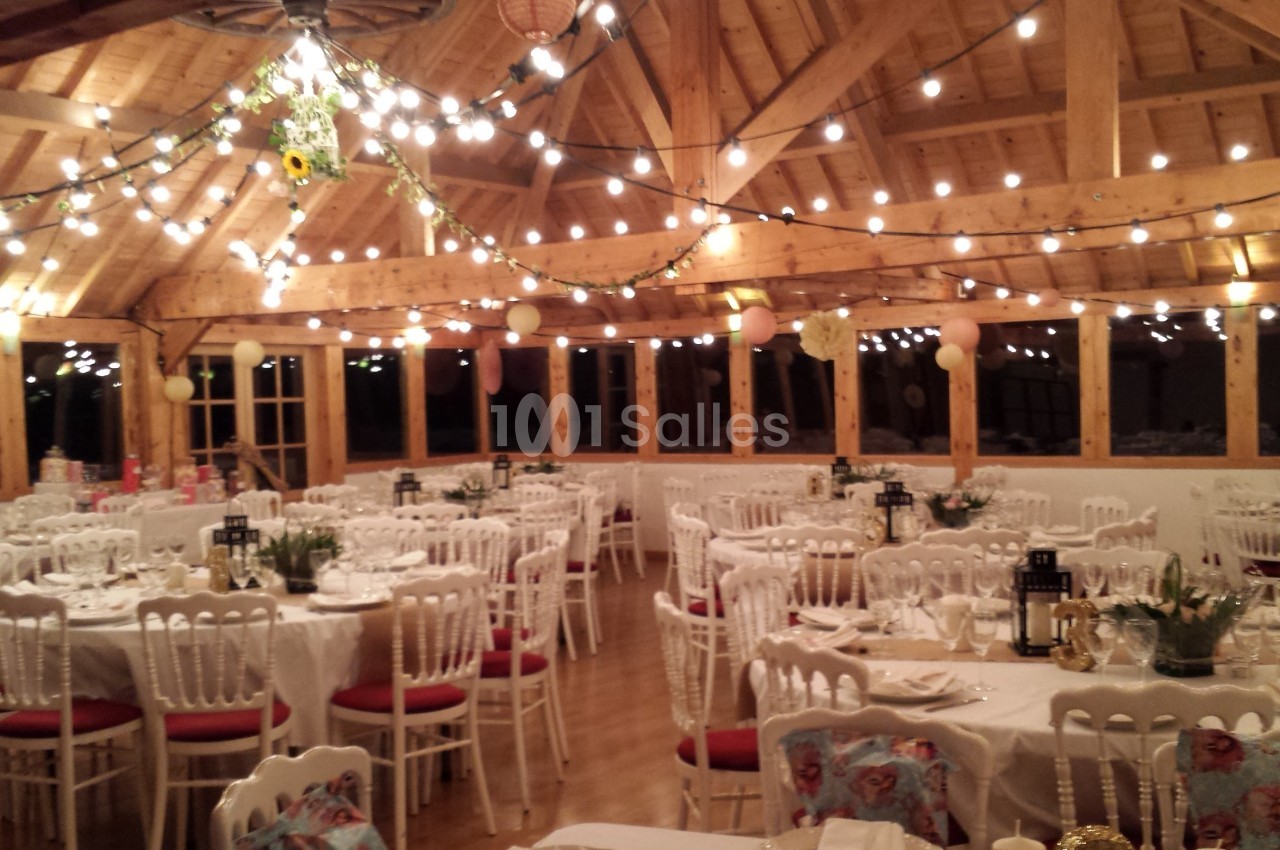 Salle de réception en bois décorée de guirlandes lumineuses, tables rondes dressées avec nappes blanches et chaises.