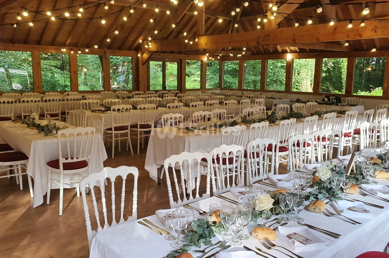 Salle de réception décorée avec des tables dressées, des chaises blanches et des guirlandes lumineuses suspendues.
