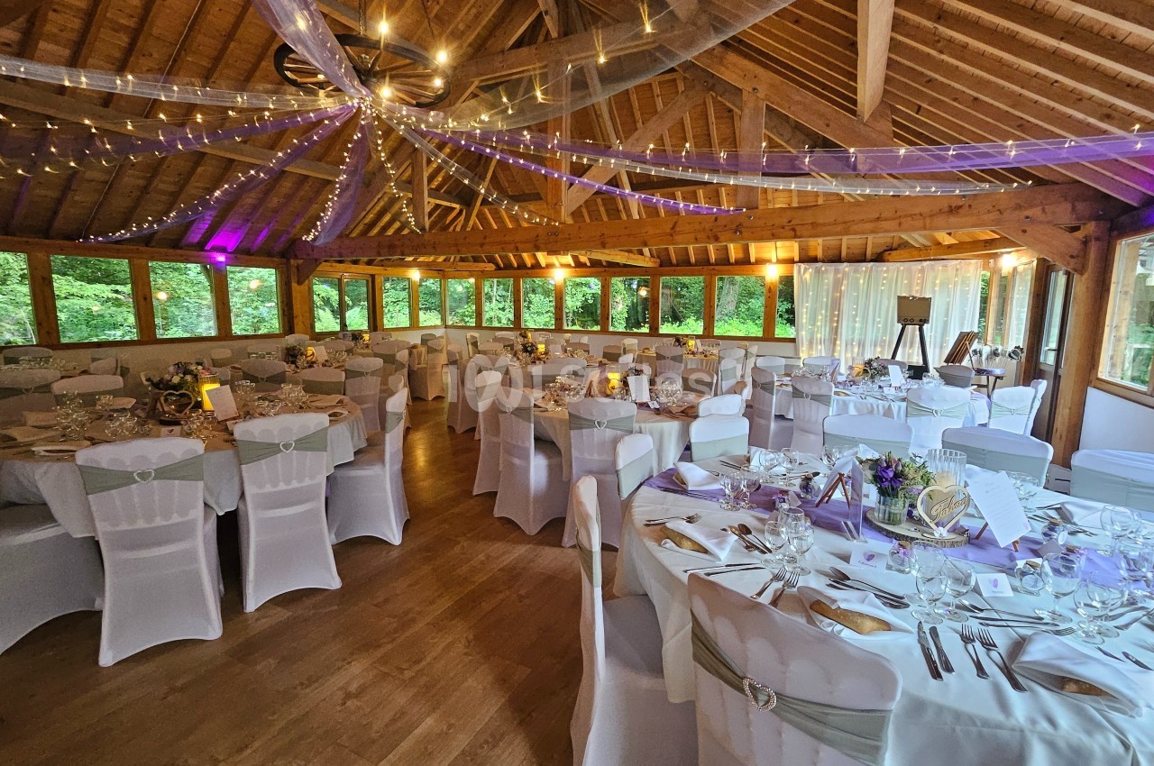 Salle de réception décorée avec des tables rondes dressées, des chaises blanches et des guirlandes lumineuses suspendues.