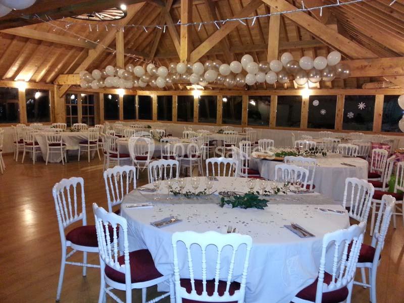 Salle décorée pour un événement avec tables rondes dressées, chaises blanches et guirlandes de ballons suspendues.