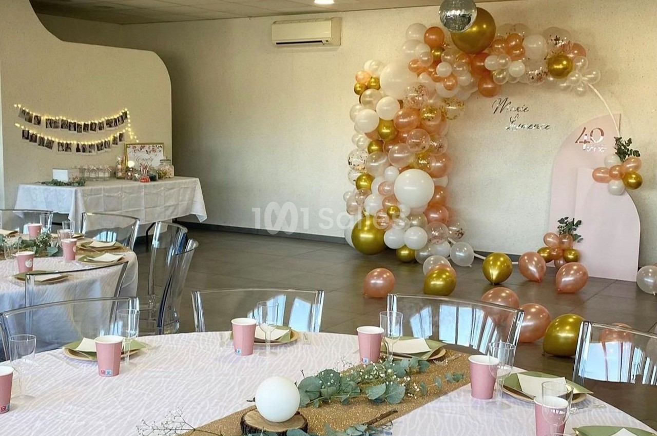 Salle décorée pour une fête avec ballons dorés et roses, tables dressées et guirlande lumineuse sur un buffet.