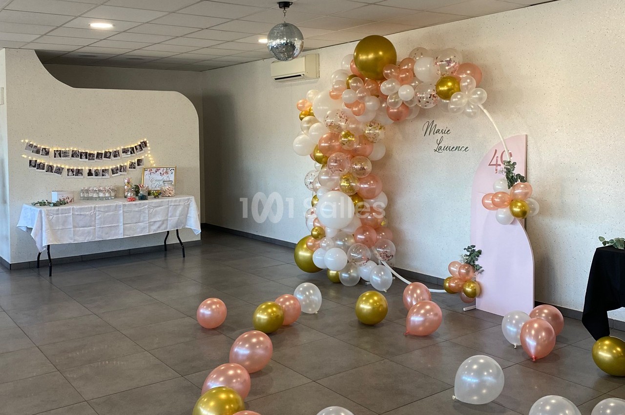 Décoration de salle avec arche de ballons roses, blancs et dorés, table décorative et guirlande de photos au mur.