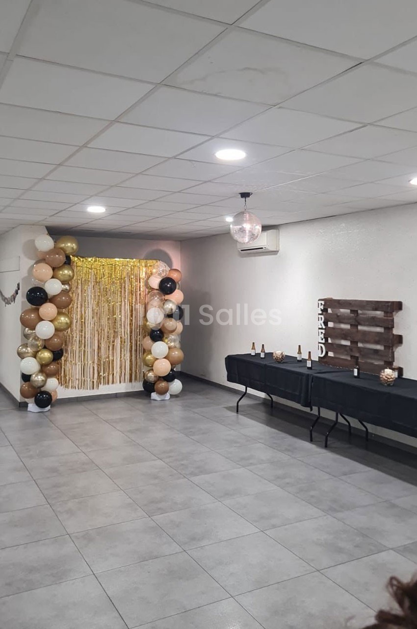 Décoration de fête avec arche de ballons beige, noir et or, rideau doré, table avec bouteilles et panneau en bois.