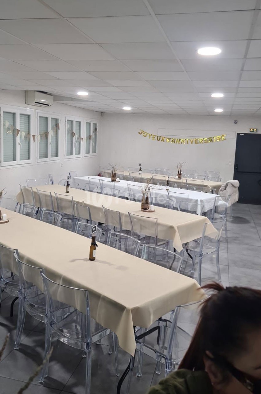 Salle décorée pour un anniversaire avec des tables alignées, nappes beiges, chaises transparentes et guirlande festive.