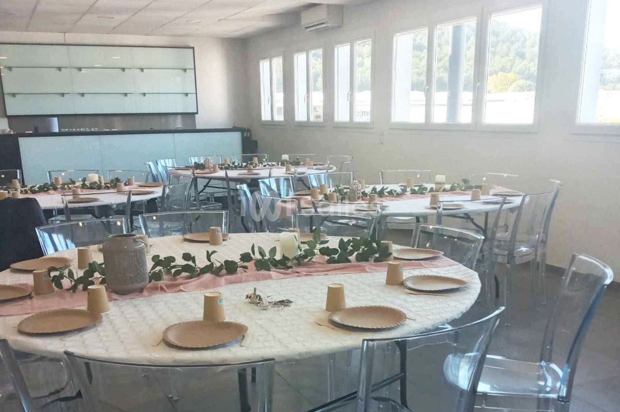 Salle lumineuse avec des tables rondes dressées pour un repas, décorées de nappes blanches et de chemins de table roses.