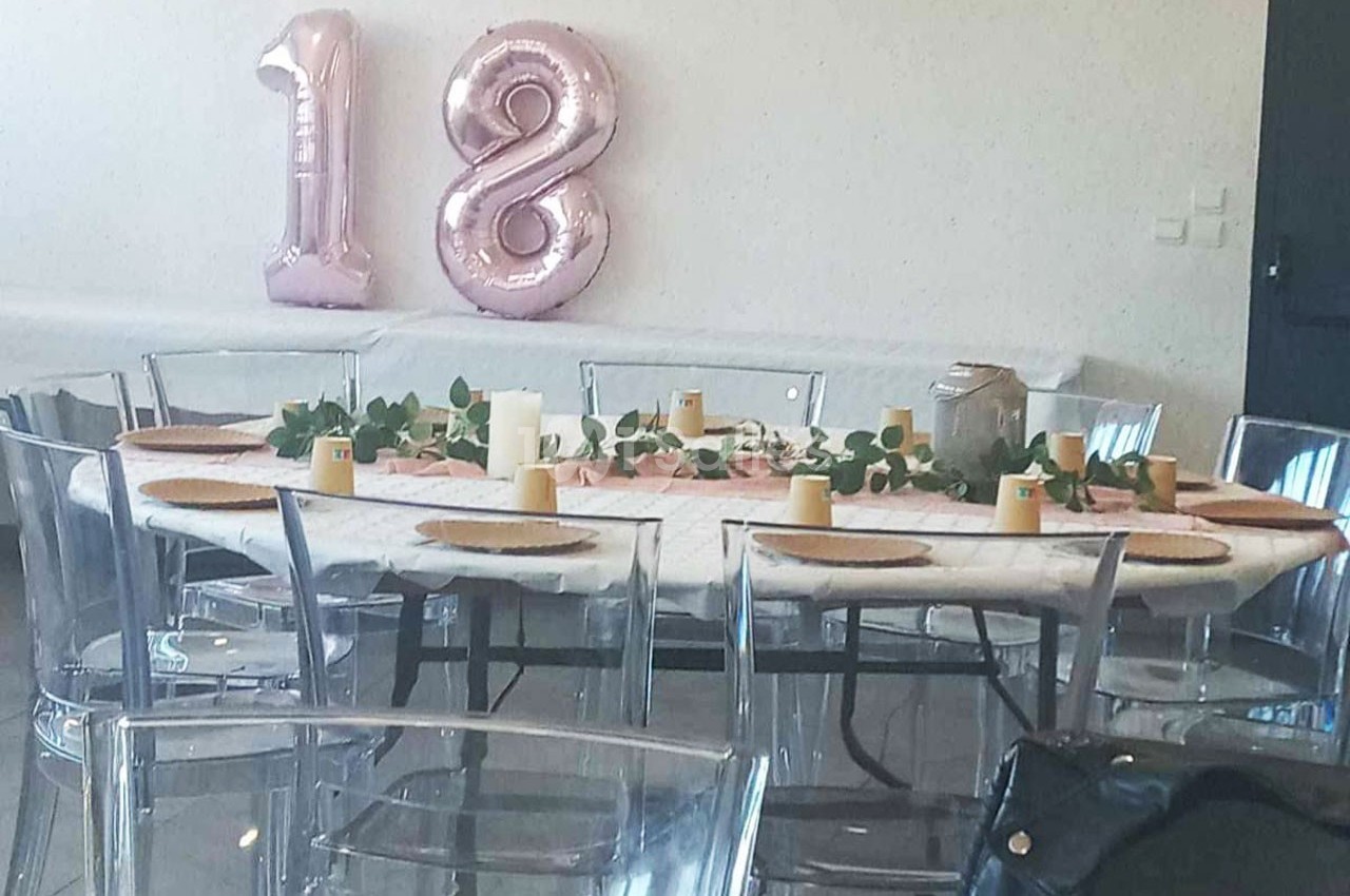 Table décorée avec des assiettes, bougies et feuillage, entourée de chaises transparentes, avec des ballons ’18’ en arrière…