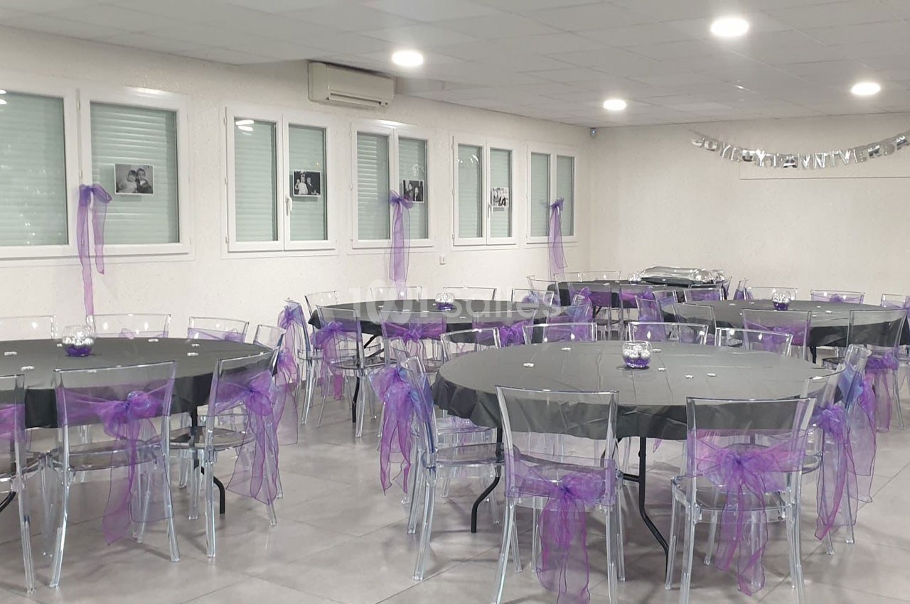 Salle décorée pour un événement avec des tables rondes nappées de noir et des chaises ornées de rubans violets.