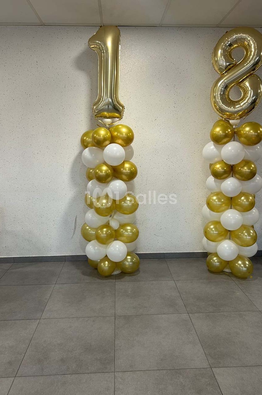 Colonnes de ballons dorés et blancs formant le chiffre 18, disposées dans une pièce au sol carrelé.