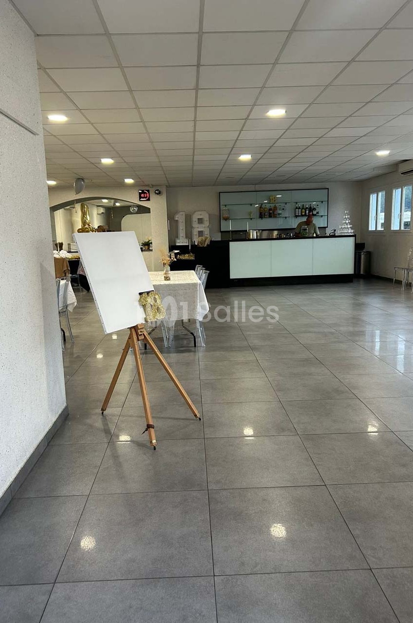 Salle lumineuse avec sol en carrelage gris, un chevalet à gauche et un comptoir de bar en arrière-plan.
