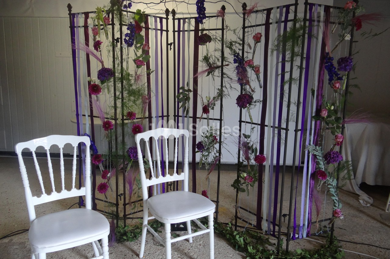 Deux chaises blanches devant une structure métallique décorée de fleurs, feuillages et rubans violets.