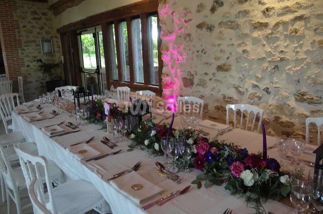 Table de réception décorée avec nappes blanches, compositions florales colorées et lanternes, dans une salle en pierre…