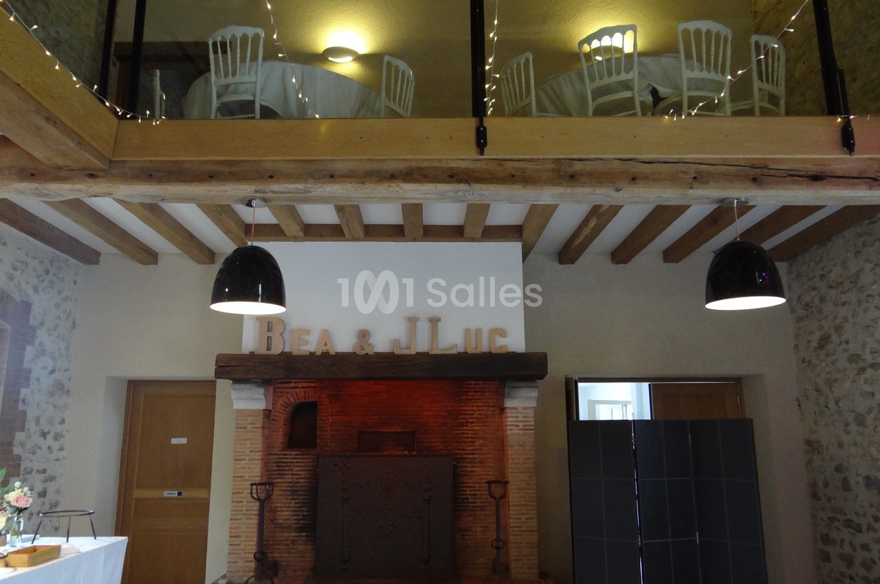 Cheminée en briques dans une salle avec poutres apparentes, tables dressées à l'étage et lettres décoratives au mur.